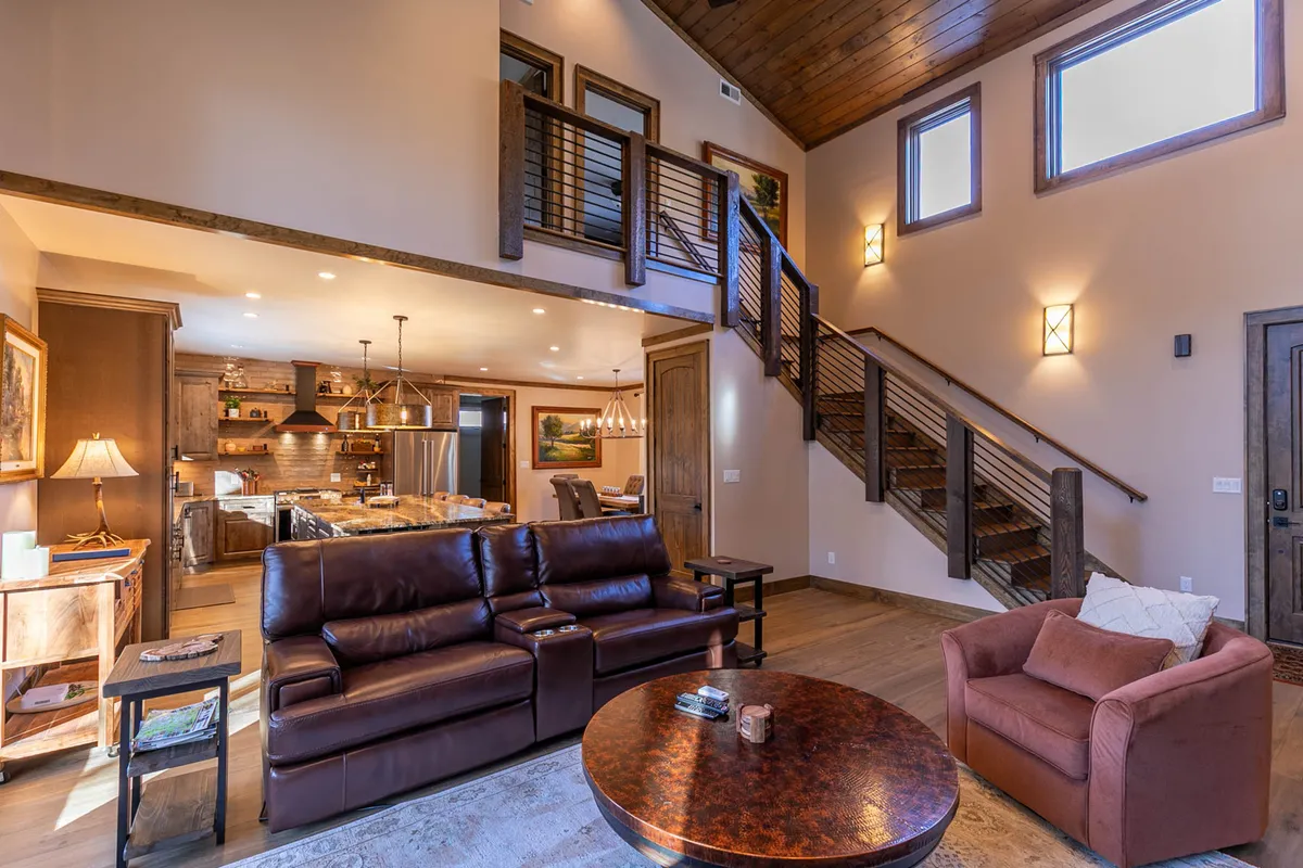 A spacious and comfortable lower level living area featuring a plush leather sectional, coffee table, and a view of the staircase and kitchen area. The arrangement suggests an inviting space for relaxing, movie nights, or sports marathons with friends and family.