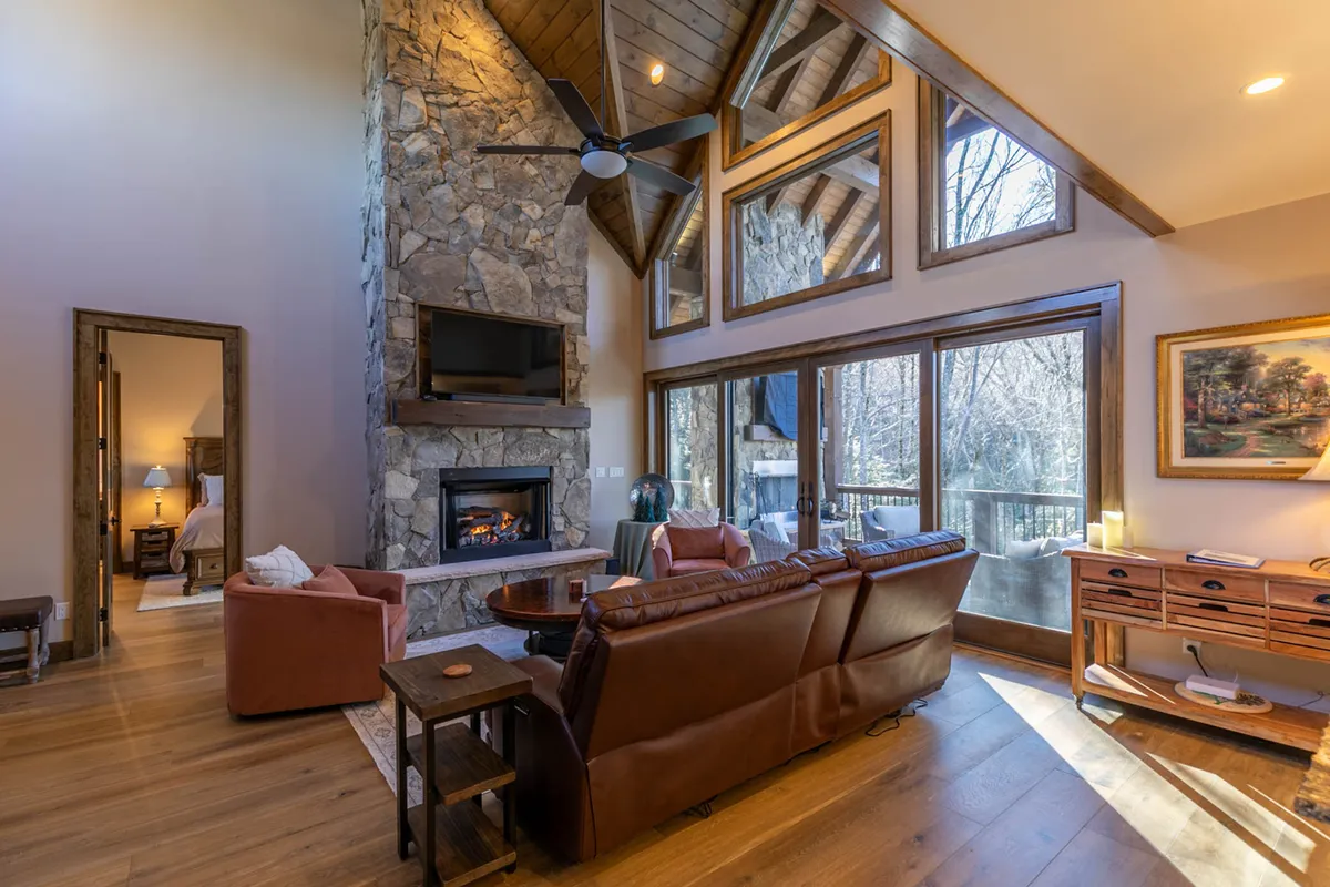 A warm, inviting living area featuring a stone fireplace, large windows that frame scenic outdoor views, comfortable seating, and a wooden table. The setting suggests a hub for social gatherings, relaxation after outdoor adventures, and cozy evenings spent together.