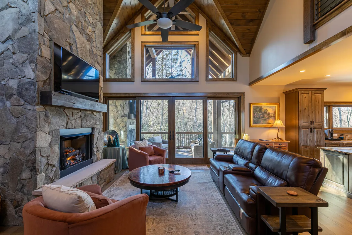 A cozy living room anchored by a dramatic floor-to-ceiling stone gas fireplace, flanked by comfortable seating and large windows that provide panoramic views of the surrounding forest. The layout invites relaxation, conversation, and game nights, embodying the heart of the lodge's communal area.