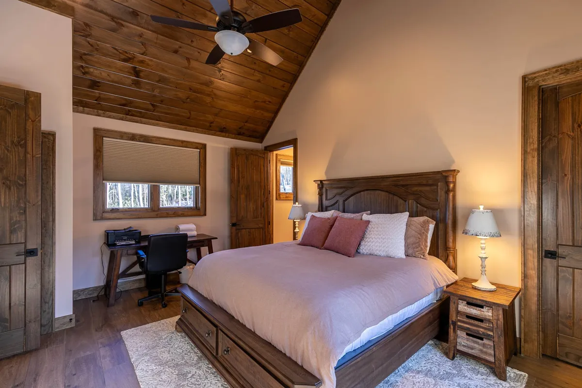 A serene interior bedroom with a desk and chair positioned under a window, offering natural light. The bed, wooden ceiling, and cozy lamp create a comfortable and practical space ideal for relaxation and work. The peaceful mountain setting is subtly visible through the window, emphasizing the connection to nature and tranquility for remote workers.