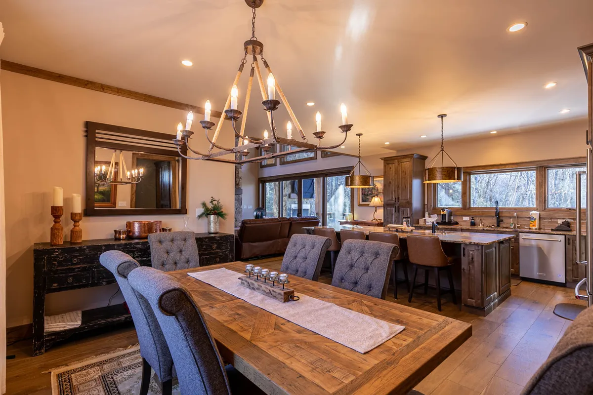 A view of the dining area featuring an expansive wooden dining table set beneath an elegant chandelier. The table is set for a large group, positioned near generous windows that flood the space with natural light. The adjacent kitchen island is visible, enhancing the sense of an open, communal space designed for group feasts and celebrations.