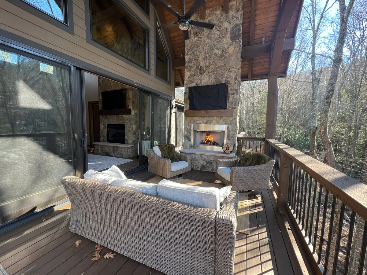 The outdoor deck, expansive and beautifully appointed, features a stone fireplace, comfortable seating arrangements, and is surrounded by lush trees. This area seamlessly extends the communal spaces outdoors, ideal for gatherings, barbecues, or tranquil mornings with coffee in hand.