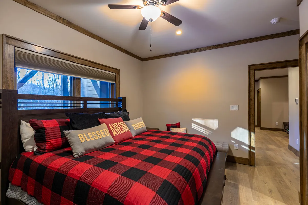 A cozy king suite bedroom featuring a wooden bed frame adorned with plaid bedding and decorative pillows. The room is enhanced by a warm ceiling fan above, creating a welcoming and restful environment perfect for relaxation, reading, or a deep night's sleep.