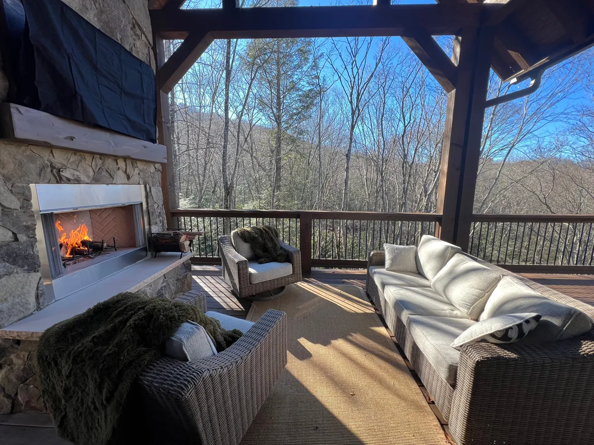 An outdoor living area features a cozy fireplace with a well-arranged seating area set amidst natural surroundings. The fire crackles as the sun sets, offering a serene space for guests to gather, socialize, or unwind with a warm drink, all while immersed in the sights and sounds of the forest.