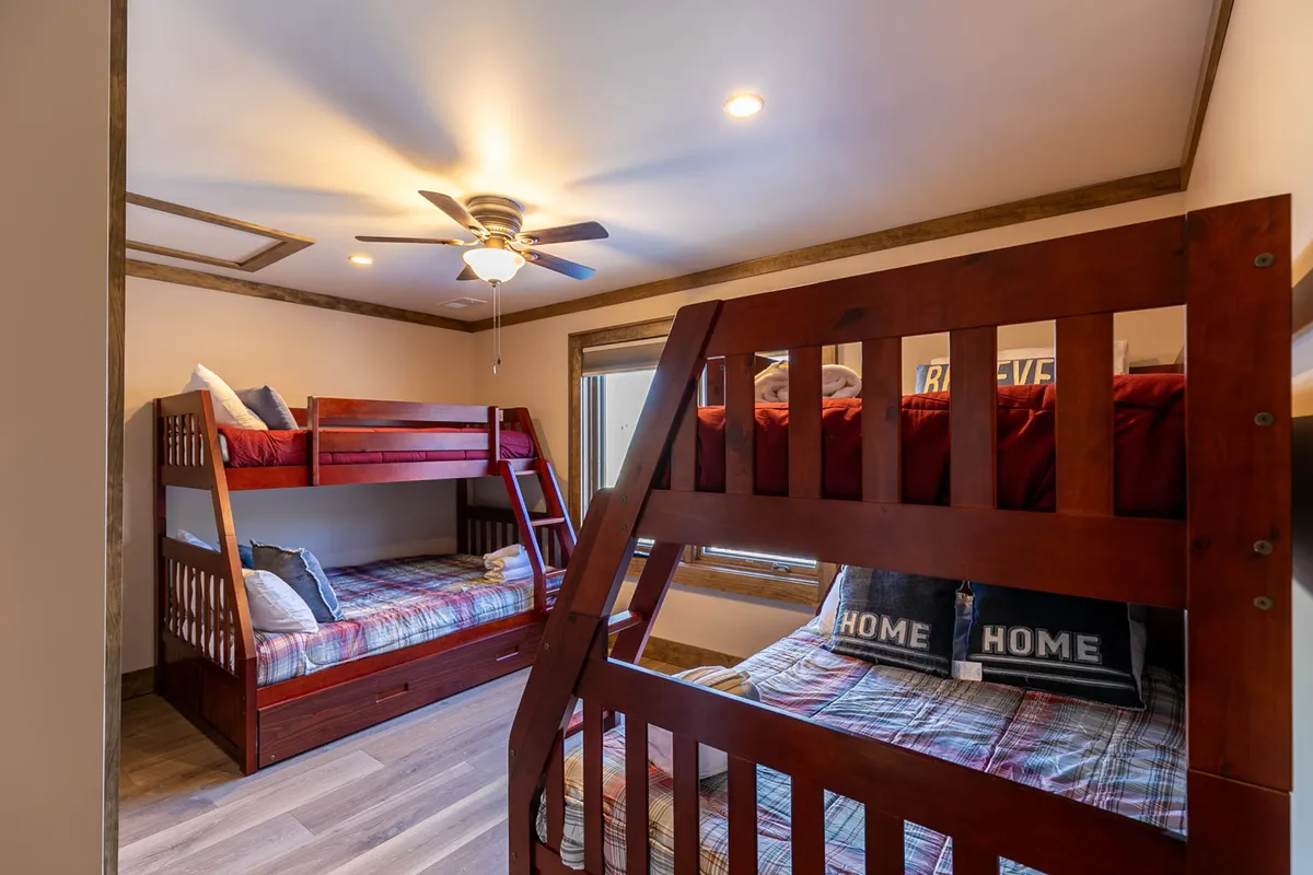 A cheerful, custom-designed bunk room with two bunk beds featuring full-size mattresses on the bottom and twins on top, plus a window allowing in natural light. The room is decorated with playful plaid bedding and a ceiling fan, making it a vibrant and welcoming space for kids or teens.