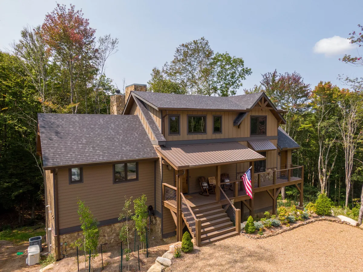 The image showcases the exterior of a beautiful single-family lodge with a welcoming front porch, classic stonework, lush landscaped garden, and an American flag waving gently. The inviting porch is perfect for sitting and taking in the tranquil mountain surroundings, while the landscaping and craftsmanship hint at the care put into this home.